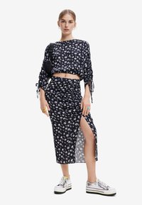 Top corto floreale nero con maniche lunghe, abbinato a una gonna midi arricciata coordinata con spacchi laterali e sneakers chunky.