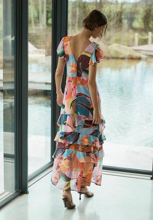 Nainen pukeutuneena värikkääseen, kerrokselliseen, kukkakuosiseen mekkoon, jossa on liehuvat hihat ja avoin selkä, seisoo suurten lasi-ikkunoiden vieressä uima-altaan luona.