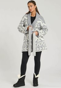 Grå och vit mönstrad stickad cardigan med bred krage, bälte och överdimensionerade ärmar, tillsammans med svarta leggings och ankelsboots.
