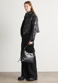 Giacca a vento nera con finitura lucida, design cropped e bottoni a pressione. La modella tiene una borsa a tracolla in pelle nera. Pantaloni larghi neri e stivali chunky.