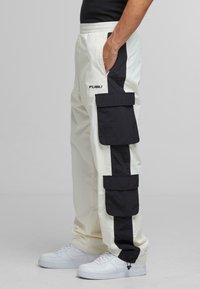White cargo pants with black side stripes and large cargo pockets. Material appears lightweight. Features elastic waistband and drawstring.