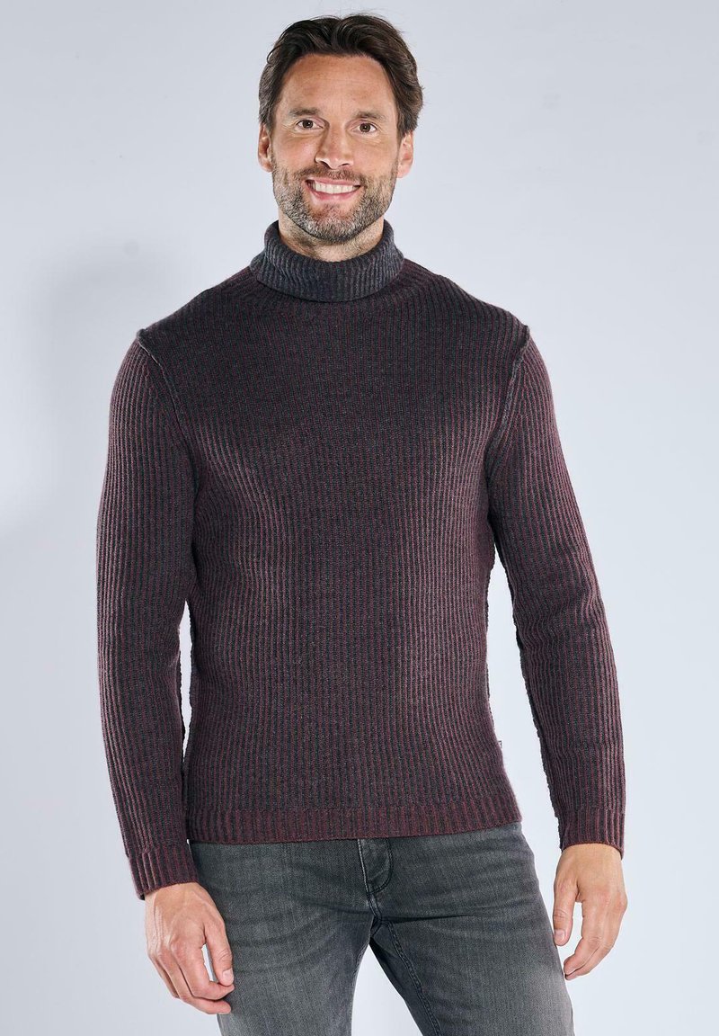 Lächelnder Mann mit kurzen braunen Haaren und Bart, der einen dunklen, gerippten Rollkragenpullover und graue Jeans trägt, vor einem einfarbigen Hintergrund.