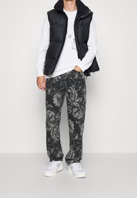 Black padded vest over a white long-sleeve shirt, paired with dark floral-patterned pants and white sneakers. Casual, layered outfit.