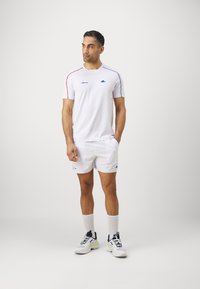 White athletic shirt with red and blue accents, paired with white shorts. Smooth texture, logo on chest, light footwear with dark accents.