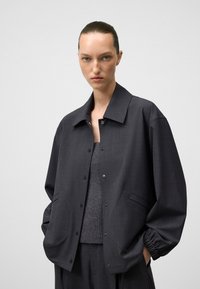 Giacca oversize grigia scura con chiusura a bottone, ampio colletto, tessuto strutturato e tasche laterali, abbinata a un maglione grigio a maglia.