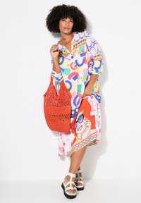 Robe colorée à motifs avec des designs géométriques et de personnages, associée à un sac en crochet rouge et des sandales chunky avec des sangles blanches.