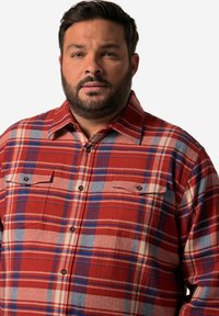 Camisa de franela a cuadros rojos y azules con cuello abotonado, dos bolsillos frontales y botones visibles, que muestra una textura suave.