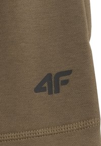 Gros plan d'un tissu olive texturé avec le logo noir "4F" et un ourlet cousu visible sur le bord inférieur.