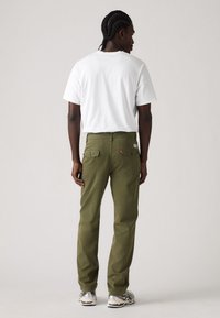 Olive green pants made of sturdy fabric with a straight leg design, featuring back pockets and a small brand label. Paired with a white t-shirt.