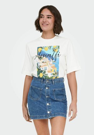 Femme souriante, portant un t-shirt blanc avec un motif de la côte Amalfitaine et une jupe en jean bleue boutonnée devant, debout devant un fond uni.