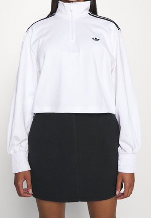 Kvinde iført en hvid kort sweatshirt med sorte striber på skuldrene og en sort mininederdel, stående foran en ensfarvet baggrund.