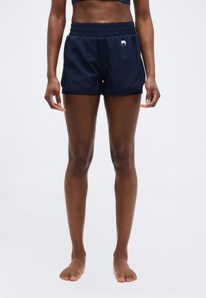 Atlétikus személy áll mezítláb, tengerészkék futóshortot viselve, bal oldalon kis fehér logóval.