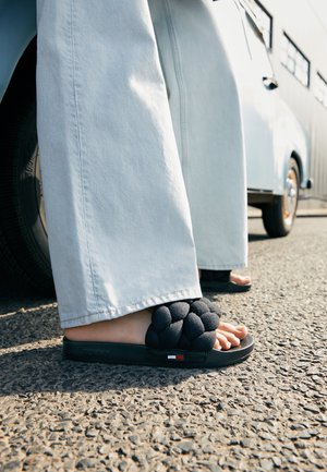 Person iført lyseblå vidde jeans og sorte flettede slides, der står på asfalt nær et hvidt køretøj.