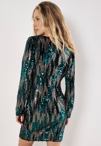 Szmaragdowa sukienka z cekinami i długimi rękawami, charakteryzująca się dopasowanym krojem i błyszczącym wzorem w pionowe linie w kolorze srebrnym i ciemnozielonym.