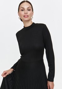 Czarna sukienka z długim rękawem, dopasowanym górnym krojem i plisowanym dołem, wykonana z gładkiego materiału. Okrągły dekolt, prosty design, bez widocznych ozdób.