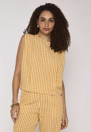Donna con capelli ricci che indossa un top senza maniche a quadretti giallo e bianco e pantaloni coordinati, in piedi davanti a uno sfondo semplice.