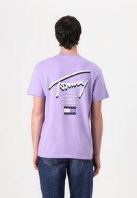 Lilla bomulls t-skjorte med et stort hvitt logo og bynavn i svart på baksiden, iført blå denimjeans.