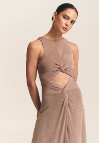 Beżowa sukienka bez rękawów, z zakręconym przodem i wycięciami. Gładka, teksturowana tkanina układa się elegancko.