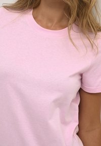 T-shirt en coton rose clair avec un col rond et des manches courtes, présentant une texture lisse et des détails de coutures minimes.