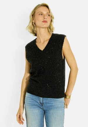 Mujer rubia con un top negro de punto sin mangas con lentejuelas y vaqueros azul claro, mirando hacia un lado con una mano detrás de la espalda.