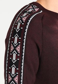 Burgundy sweater featuring a patterned black and white knit design along the shoulder, with geometric accents in red and white.