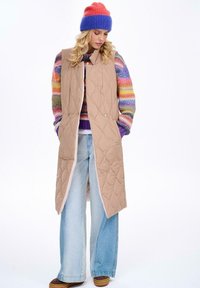 Beżowa, pikowana długa kamizelka z kołnierzem, zapinana na zatrzaski, noszona na wielokolorowym swetrze dzianym i jasnoniebieskich, szerokich dżinsach.