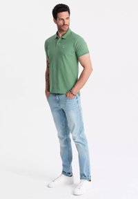Camisa polo verde de manga corta con un logo sutil, combinada con jeans de color azul claro y zapatillas blancas, de pie contra un fondo liso.