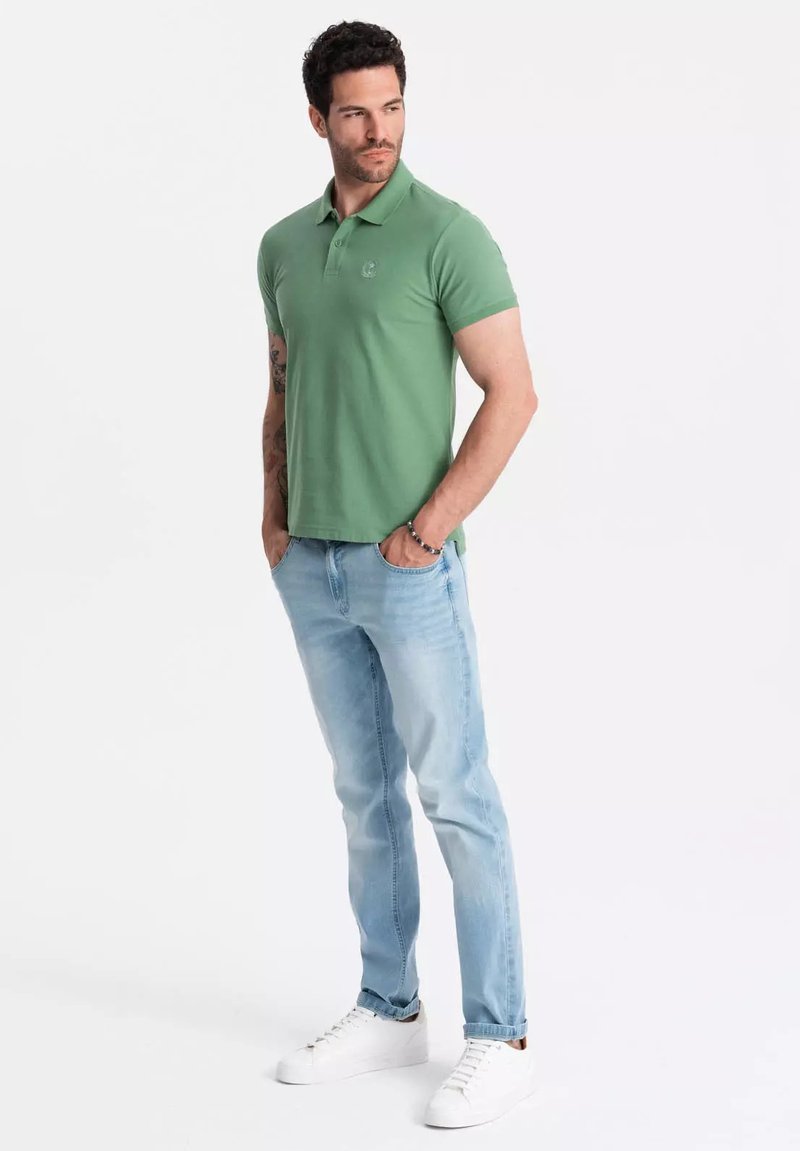Camisa polo verde de manga corta con un logo sutil, combinada con jeans de color azul claro y zapatillas blancas, de pie contra un fondo liso.