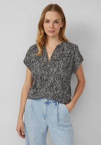 Schwarz-weiß gemusterte Bluse mit V-Ausschnitt, kurzen Ärmeln, lockerer Passform und geometrischen Designs; kombiniert mit hellblauen Jeans.