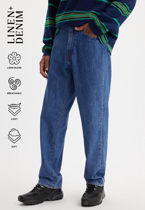 Homme portant un jean large en mélange lin bleu avec des baskets noires et une chemise à manches longues rayée, debout avec une main dans la poche.