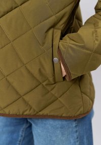 Olivzaļš pūkains jaciņa ar diagonālu šuvi, kontrastējošām brūnām apmalēm, sānu kabatu ar spiedpogu un samtainu auduma faktūru.