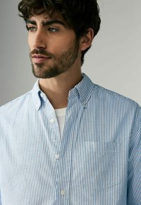 Chemise rayée bleu clair et blanche avec un col boutonné, une poche avant et une texture de tissu douce. Portée par-dessus un T-shirt blanc.