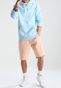 Sweat à capuche bleu clair avec poche avant, associé à un short pêche. Des baskets blanches complètent la tenue. Design simple et décontracté.