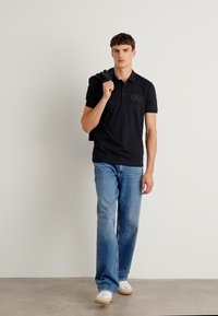 Camisa polo preta com um pequeno logotipo, combinada com jeans azul. O modelo leva um suéter azul-marinho sobre o ombro, usando tênis brancos.