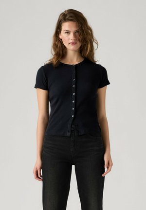 Jonge vrouw met golvend bruin haar, een nauwsluitende zwarte korte mouw knoopblouse en zwarte broek tegen een egaal lichte achtergrond.