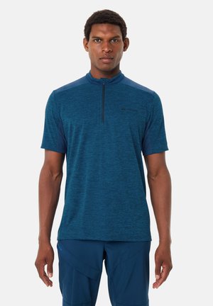 Uomo in piedi di fronte, indossa una maglietta blu con zip a maniche corte e pantaloni blu coordinati, davanti a uno sfondo bianco neutro.