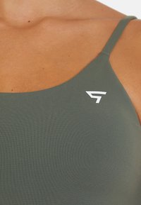 Sujetador deportivo verde oliva con tirantes finos, que presenta una tela texturizada y un pequeño logotipo blanco en el lado izquierdo.
