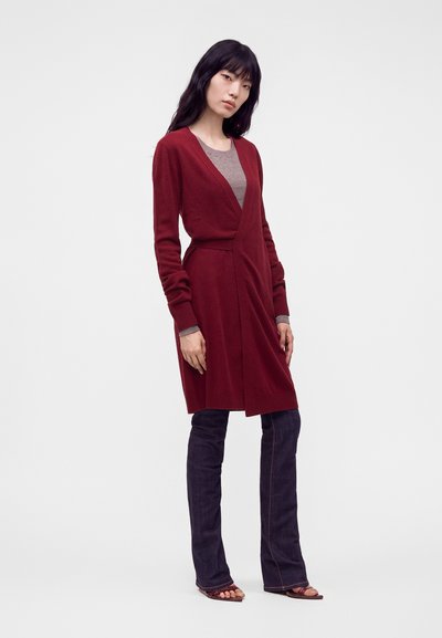 Femme aux longs cheveux foncés portant un long cardigan bordeaux, un haut gris, un jean foncé et des sandales marron, debout devant un fond clair uni.