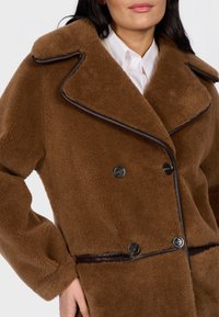 Donna che indossa un cappotto in finta shearling marrone a doppio petto con profili scuri sopra una camicia bianca con colletto, mano poggiata in tasca.