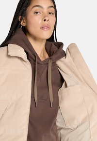 Giacca in corduroy beige con zip, tasche interne, abbinata a una felpa marrone con cappuccio, dotata di lacci e logo.