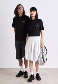 Dos personas vistiendo camisetas polo negras con un logo blanco, una lleva pantalones cortos negros y gafas de sol, y la otra una falda plisada blanca sosteniendo un bolso plateado.