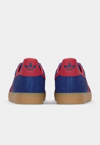 Et par blå og røde Adidas-sneakers med gummisåler, set fra bagfra, der viser Adidas-logoet på hælflapperne.