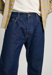 Jeans de mezclilla azul oscuro con un corte clásico, con costuras visibles y un cierre frontal con botón. Combinados con una camisa de cuadros amarilla y gris.