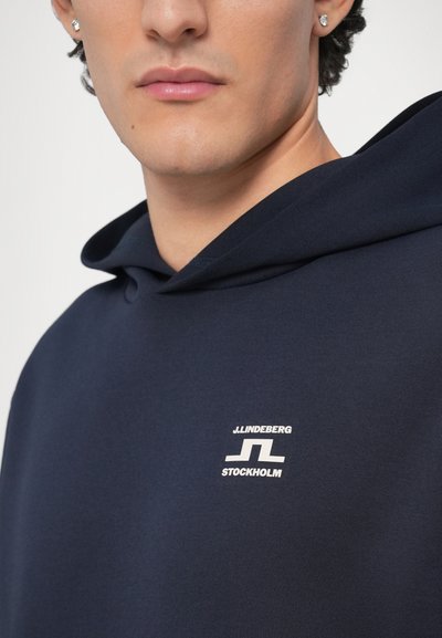 Μπλε ναυτικό φούτερ με κουκούλα από απαλό ύφασμα, με λευκό λογότυπο στο στήθος. Δομημένη λαιμόκοψη και λεία υφή χωρίς επιπλέον μοτίβα.