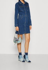 Jeansklänning med långa ärmar, knappstängning, två bröstfickor och ett knutet bälte. Matchad med en iriserande väska och blå klackstövlar med blockklack.