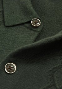 Material tricot de culoare verde închis, cu două butoane cu margini metalice și o detaliere a gulerului pliat vizibilă.