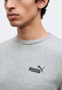 Gros plan sur une personne portant un sweatshirt Puma gris clair avec le logo Puma noir sur la poitrine.