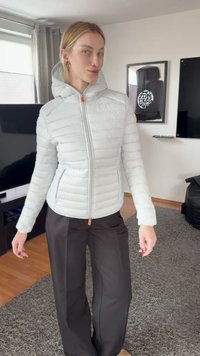 Γυναίκα με ανοιχτό γκρι επενδυμένο μπουφάν και σκούρο παντελόνι φαρδύ στο πόδι, στέκεται σε σαλόνι με τηλεόραση και κορνιζαρισμένα έργα τέχνης στον τοίχο.