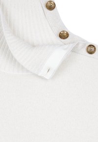 Tissu tricoté côtelé blanc avec trois boutons dorés décoratifs présentant un motif de crâne et de bouclier le long d'un bord plié.