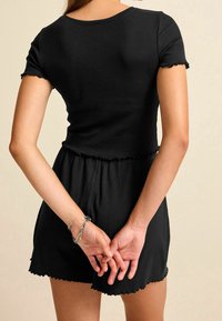 Vrouw in een zwart korte mouwen jurk met rimpelrand, met haar rug naar de camera gekeerd, handen gevouwen achter haar rug, met een zilveren armband.